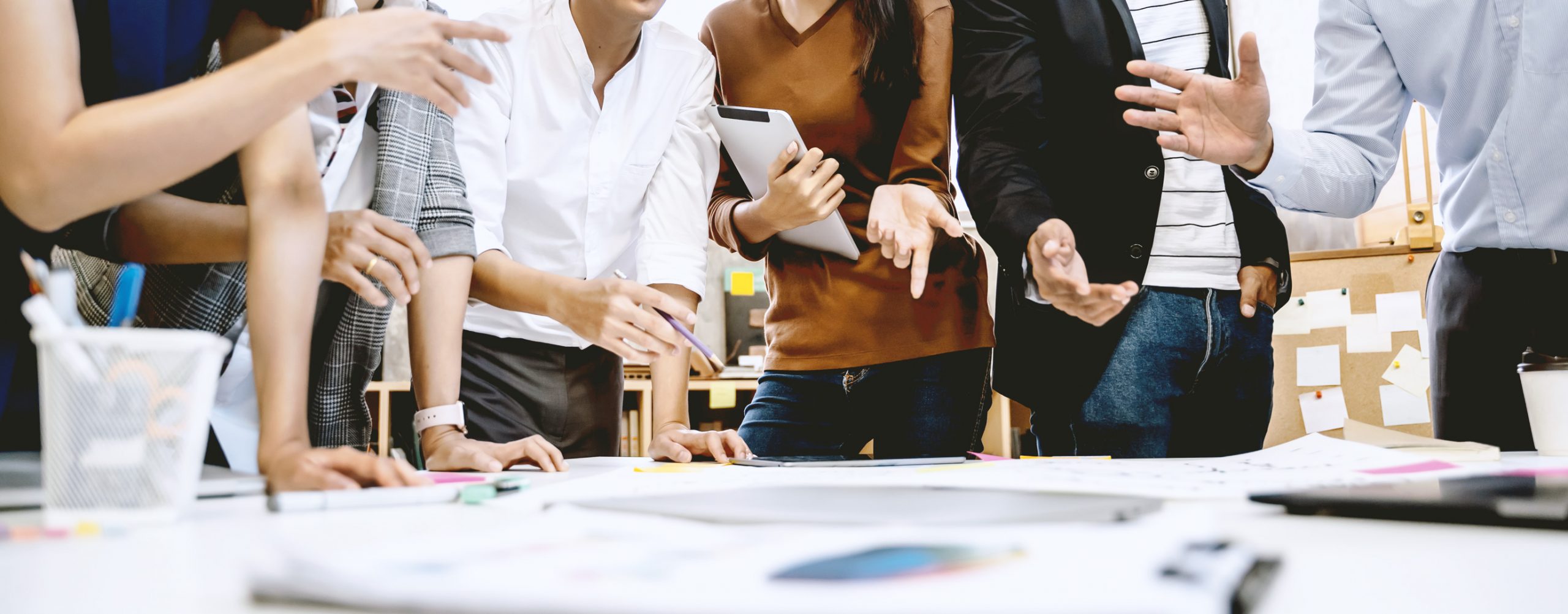 Team-Besprechung am Tisch. Der Bildausschnitt zeigt die Oberkörper mehrerer Personen, die um einen Tisch stehen oder sich darauf abstützen. Auf der Tischfläche liegen Dokumente, auf die die Hände der Teilnehmenden gestikulierend deuten. Die Gesichter sind nicht im Bild.