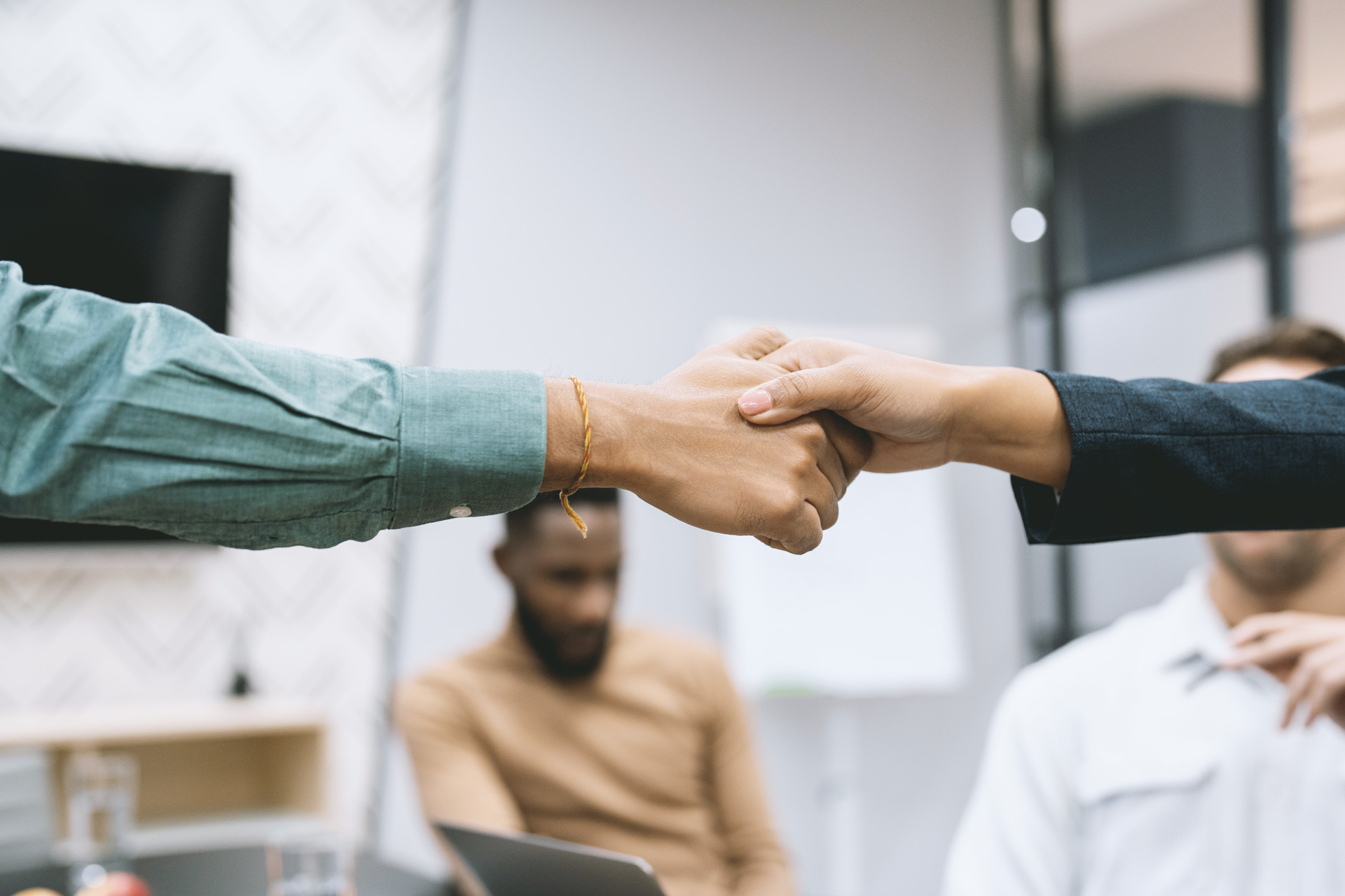 Ein Handschlag. Zwei Hände geben sich die Hand. Im Hintergrund sitzen zwei Personen.