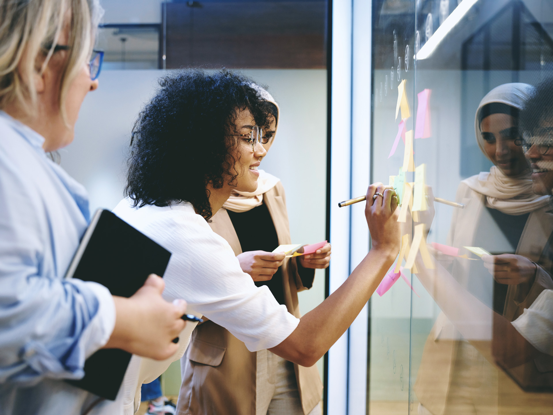 Drei Frauen stehen vor einer Glaswand mit Post-its. Eine beugt sich vor und schreibt auf einen Zettel, die zweite hält weitere Post-its in der Hand, die dritte beobachtet die Szene.